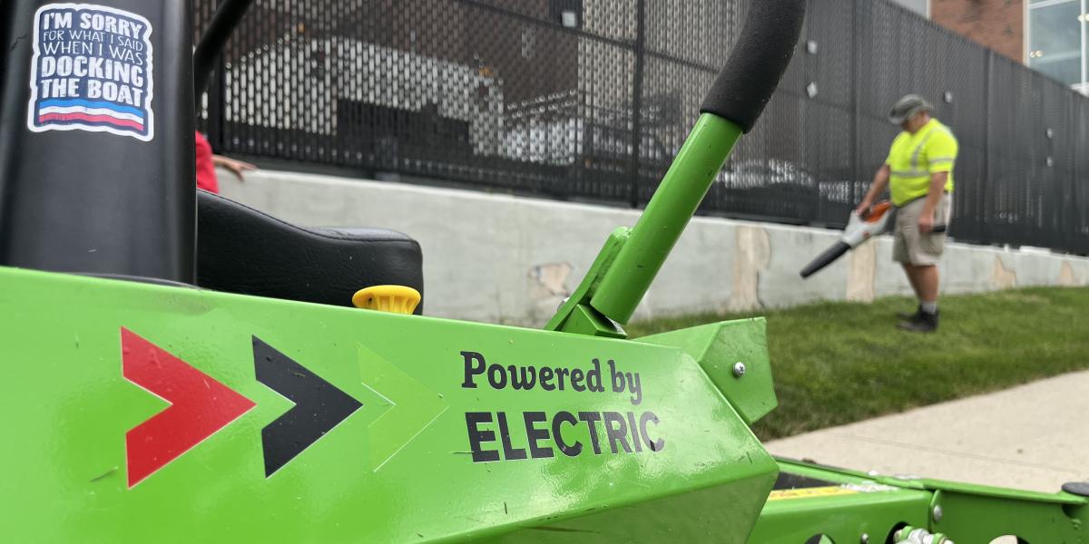 An electric mower with a landscaper in the background cleaning up grass.