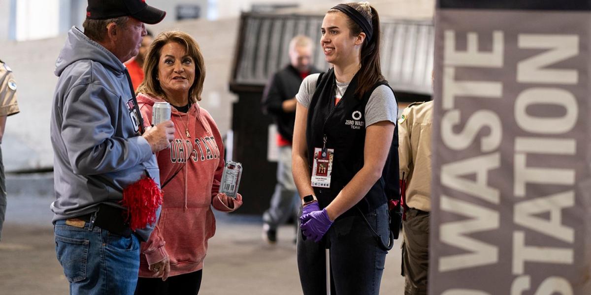 Zero waste volunteer chats with man and woman near recylcing bins inside stadium