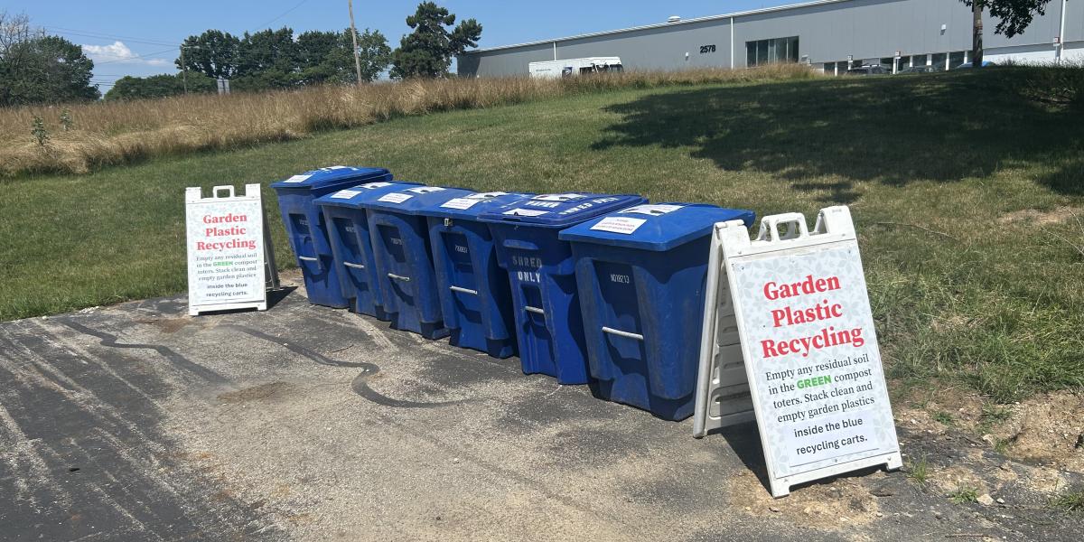 Six blue recycling bins are seen with an A-frame sign on each side about recycling. 