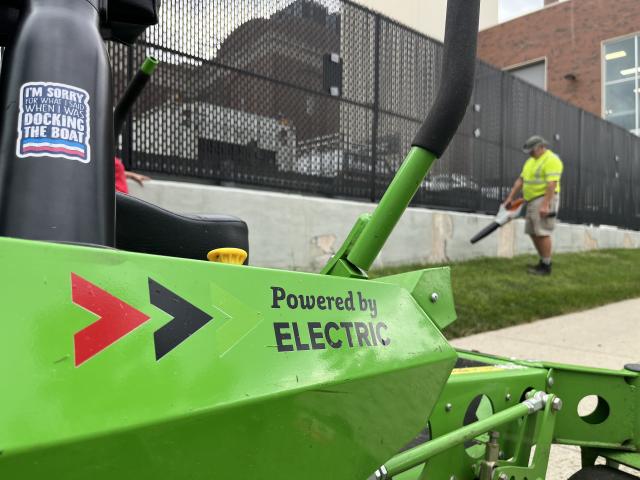 An electric mower with a landscaper in the background cleaning up grass.