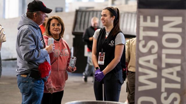 Zero waste volunteer chats with man and woman near recylcing bins inside stadium