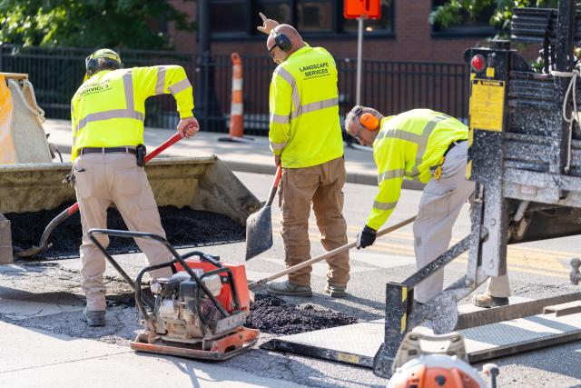 Three men seen wearing bright yellow 'Landscape Services' shirts working on fixing a road.