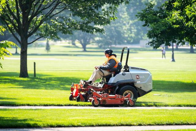 A man seen riding a lawnmower in a large field with trees.