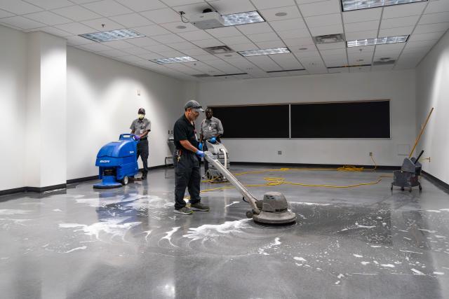 Three men seen using equipment to clean hard, gray floors in a classroom.